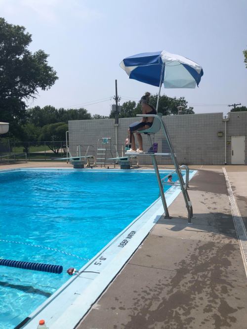 A lifeguard sits on a tall chair under an umbrella, overlooking a clear blue swimming pool on a sunny day.