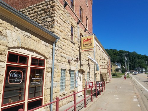 Historic stone building with a sign for "Potosi Brew Pub," featuring large windows and a staircase outside. Clear blue sky.