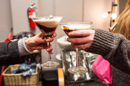 Two hands holding cocktails in elegant glasses, one with a layered chocolate and cream drink, the other a creamy beverage.