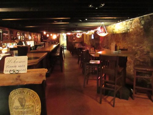 A dimly lit bar interior with wooden tables, chairs, and a long counter, featuring warm lighting and stone walls.
