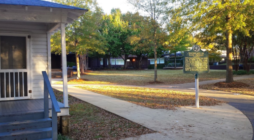 A pathway leads through a park with trees, near a historical marker and a white house in the foreground.