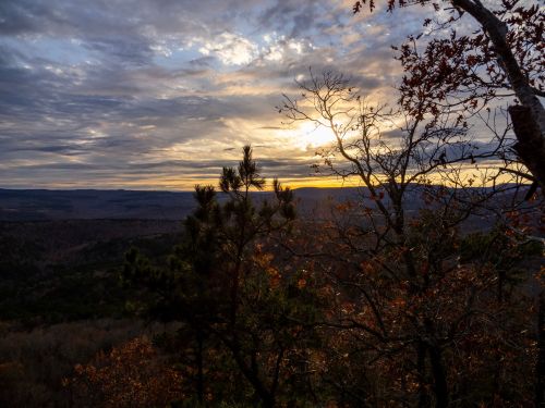 Sunset over a mountainous landscape, with trees in the foreground and a dramatic sky filled with clouds.