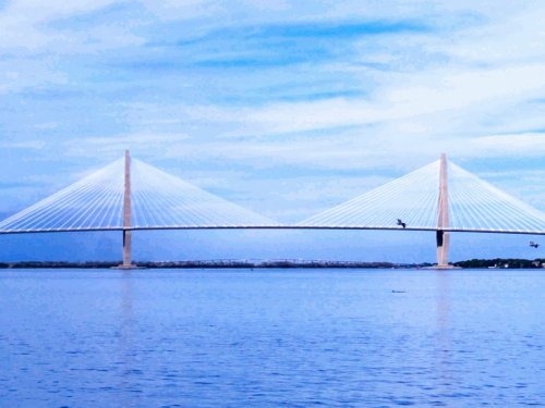 A modern suspension bridge spans a calm body of water under a blue sky with wispy clouds.