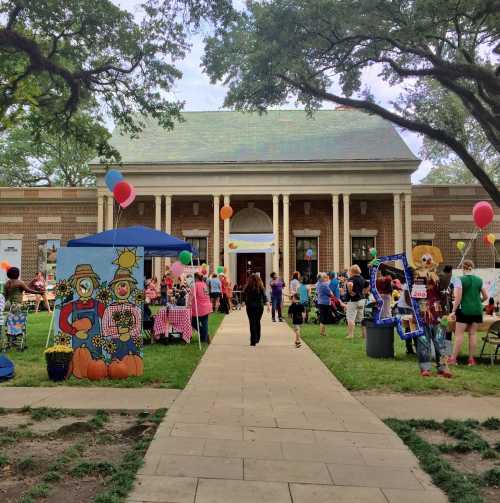 A lively outdoor event with people, colorful decorations, and balloons in front of a brick building.