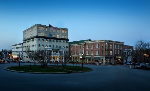 Historic buildings at dusk, featuring a modern hotel and a brick structure, with trees and streetlights in the foreground.