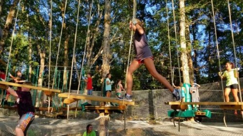 A group of children and teens navigate a ropes course in a wooded area, balancing on wooden beams and enjoying the activity.