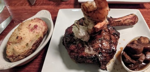 A plate featuring a grilled steak topped with shrimp, a baked potato, and a side of mushrooms.
