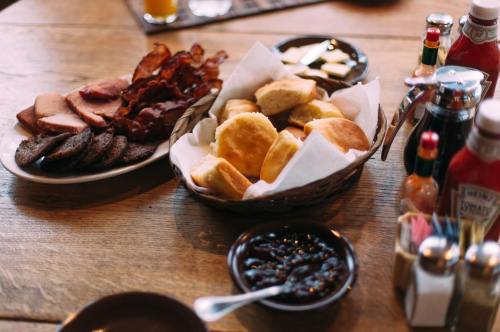 A wooden table with a basket of biscuits, a plate of bacon and ham, and condiments like ketchup and jelly.