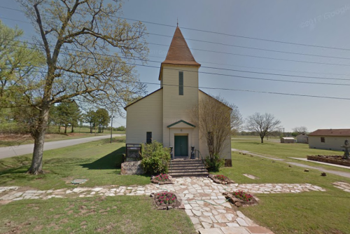 A small, rustic church with a steeple, surrounded by greenery and a stone pathway, under a clear blue sky.