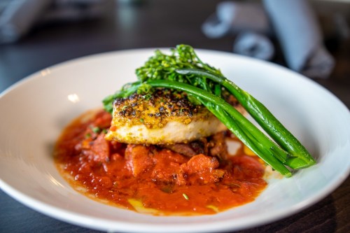 A plate of fish topped with herbs, served on a bed of tomato sauce and garnished with green vegetables.