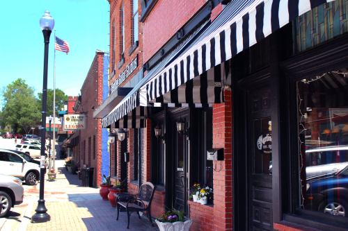 A charming street scene featuring brick buildings with striped awnings, flower pots, and parked cars under a clear blue sky.
