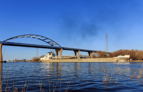 A large ship passes under a modern bridge, reflecting in calm water with clear blue skies above.