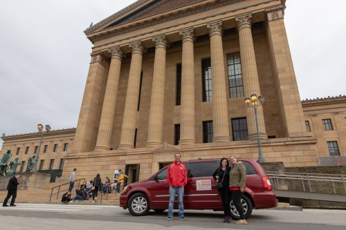 A red minivan parked in front of a large, classical building with columns, two people stand beside it.
