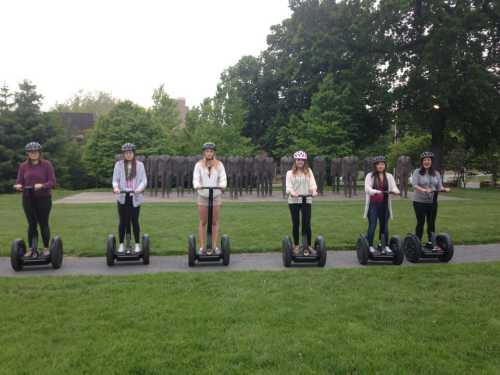 A group of six women wearing helmets stand on Segways in a park, surrounded by greenery and trees.