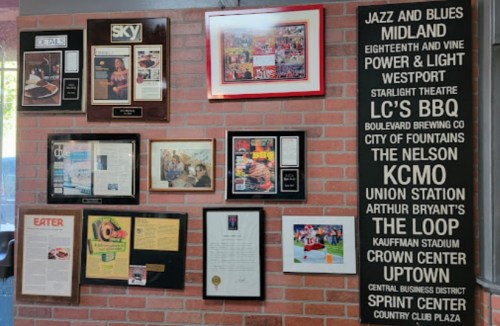 A wall display featuring framed awards, articles, and a list of local venues and attractions in a restaurant setting.