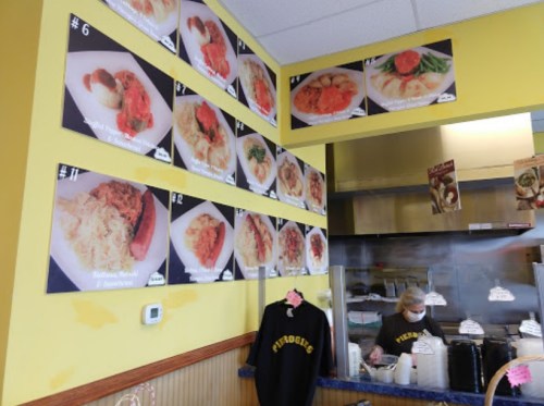 Menu wall featuring various dishes at a restaurant, with a staff member in the foreground wearing a black sweatshirt.