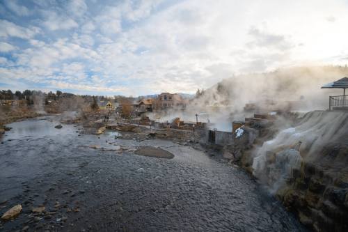 Steam rises from hot springs along a river, with buildings and trees in the background under a cloudy sky.