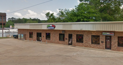 A brick building with multiple storefronts, featuring signs for food and entertainment, surrounded by trees and a parking lot.