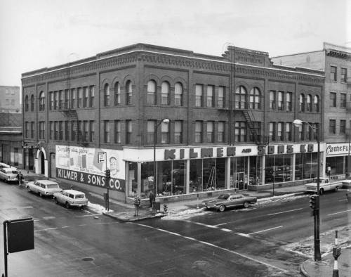 Historic black-and-white photo of a brick building with "Kilmer & Sons Co." signage, located at a street corner.