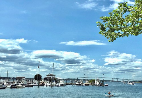 A scenic view of a harbor with boats, under a blue sky filled with clouds, and a bridge in the background.