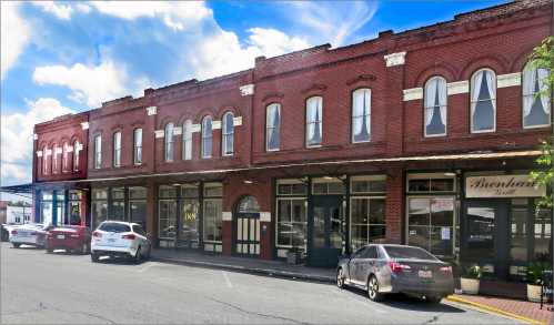 Historic brick buildings line a street, featuring shops and parked cars under a bright blue sky.