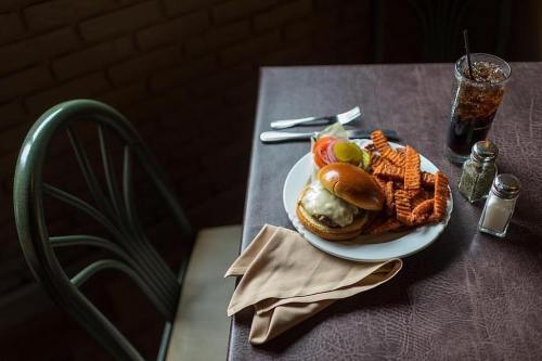 A plate with a cheeseburger, lettuce, tomato, pickles, and sweet potato fries, next to a glass of iced drink on a table.