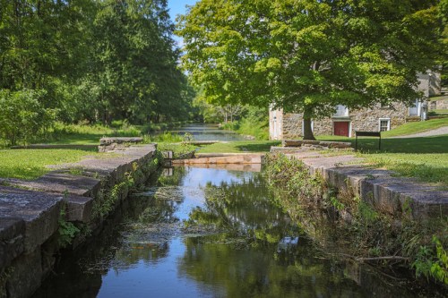A serene landscape featuring a calm stream, lush greenery, and a stone building in the background.
