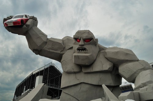 A giant stone statue of a monster holding a car, set against a cloudy sky.