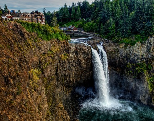 A waterfall cascades down rocky cliffs, surrounded by lush greenery and a distant lodge in a scenic landscape.
