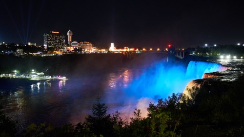 Niagara Falls illuminated at night with colorful lights, surrounded by city buildings and mist.