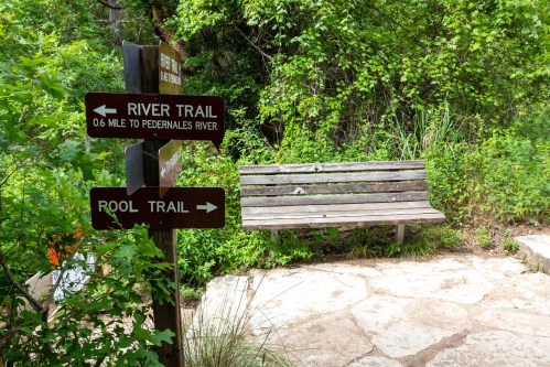 A wooden bench near a trail sign pointing to River Trail and Pool Trail, surrounded by lush greenery.