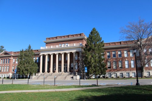 A large brick building with columns, surrounded by trees and a clear blue sky, set in a grassy area.