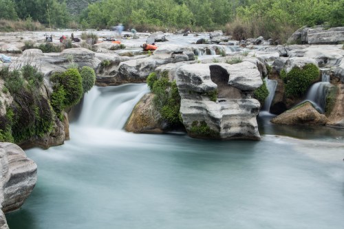 A serene river flows over rocky formations, surrounded by lush greenery and kayakers in the distance.