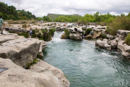 A serene river flows through rocky terrain, surrounded by lush greenery and cloudy skies. People explore the area.