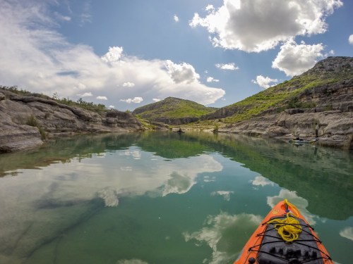 A kayak on calm water reflects clouds and mountains under a blue sky, surrounded by lush greenery.