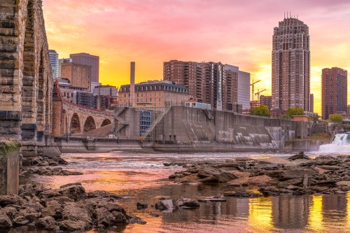 A vibrant sunset over a city skyline, with a river and rocky shore in the foreground, showcasing urban architecture.