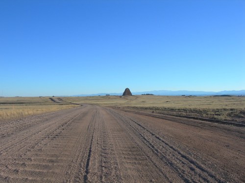 A dirt road stretches into the distance, leading to a distant hill under a clear blue sky.