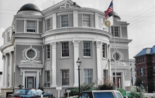 Historic building with a rounded facade, flagpole, and decorative elements, surrounded by greenery and parked cars.