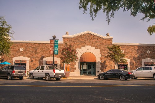 A brick building with an arched entrance, surrounded by trees and parked cars, featuring colorful banners.
