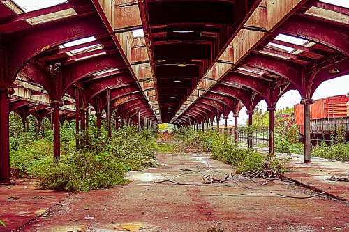 Abandoned train station with overgrown plants, rusted metal beams, and a long, empty platform under a cloudy sky.
