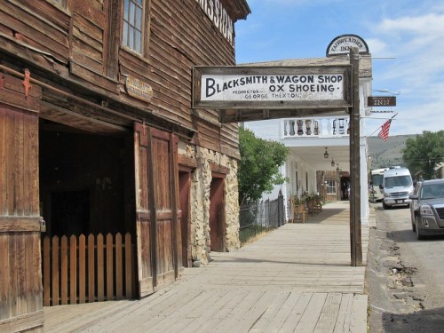 A rustic street scene featuring a blacksmith and wagon shop sign, with wooden buildings and a glimpse of parked vehicles.