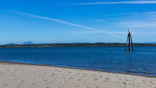 A serene beach scene with calm water, a distant mountain range, and a wooden post marking the shoreline under a clear blue sky.
