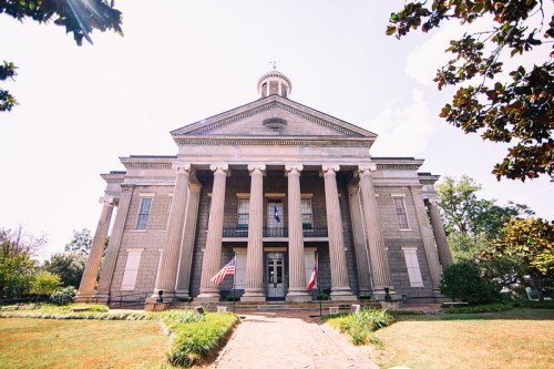 Historic building with tall columns, flags, and a dome, surrounded by greenery under a clear blue sky.