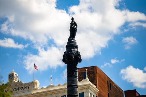 A tall monument topped with a statue, set against a blue sky with clouds and a historic building in the background.