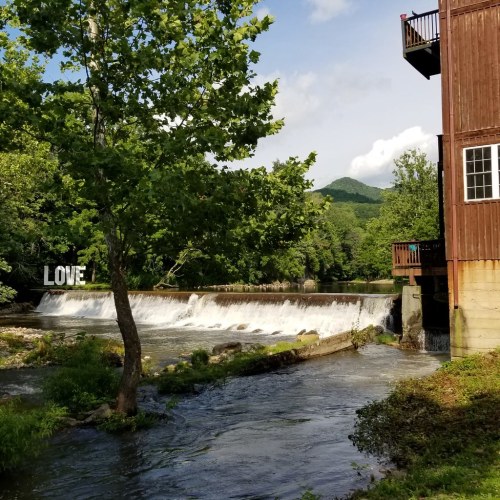 A serene river with a waterfall, surrounded by trees and a wooden building, under a blue sky with clouds.