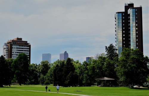 A park scene with two people walking a dog, surrounded by trees and tall buildings in the background under a cloudy sky.