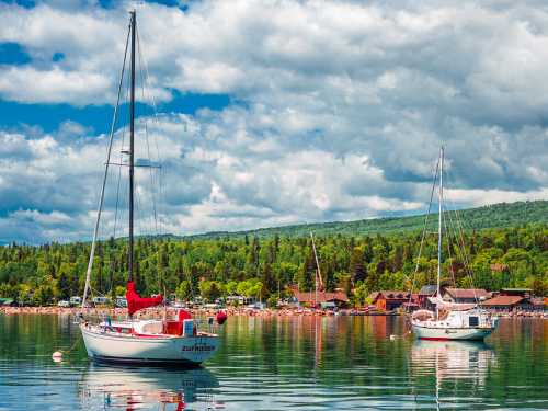 Two sailboats anchored in a calm lake, surrounded by lush green trees and a cloudy blue sky.