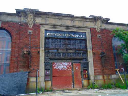 Abandoned brick building with a sign reading "Pawtucket-Central Falls," featuring boarded windows and graffiti.