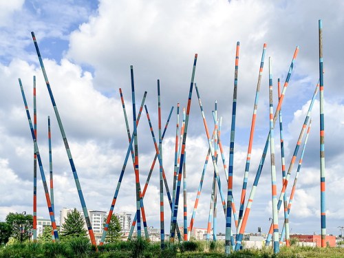 Colorful tall poles in a grassy area under a cloudy sky, with buildings visible in the background.