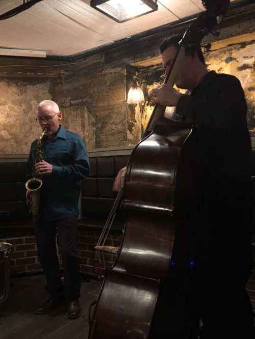 Two musicians perform in a dimly lit venue; one plays the saxophone while the other plays the double bass.